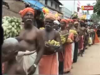 A procession of people in orange and white sarongs carries offerings. The Vasanth TV logo sits in the corner as they move down the street in India.
