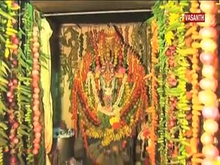 A deity, garlanded in vibrant flowers, fills the central space. Strings of colorful decorations hang on either side, framing the scene, with the Vasanth TV logo in the corner.
