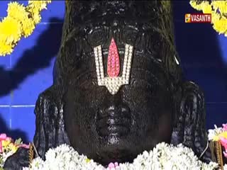 A dark, sculpted figure, adorned with a jeweled emblem, is framed by yellow and white flowers. The Vasanth TV logo sits in the top right corner.
