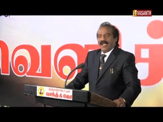A man in a dark suit stands at a podium, speaking into a microphone. The Vasanth TV logo is visible in the corner, and a large graphic is behind him.
