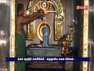 A man in a green cloth pours liquid from a vessel onto a dark, cylindrical stone. The scene is framed by ornate gold details, with the Vasanth TV logo visible in the corner.

