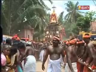 A procession of people, many shirtless and wearing turbans, moves forward, carrying a decorated structure. The Vasanth TV logo is visible as the crowd advances through a sunny, tree-lined street in India.

