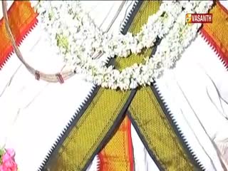 A white garland of flowers hangs over a traditional white garment with gold and red trim. The Vasanth TV logo is visible in the upper right corner.
