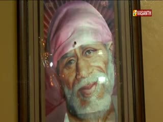 A framed portrait of a man with a white beard and a pink head covering hangs on the wall. The Vasanth TV logo is visible in the corner of the frame.
