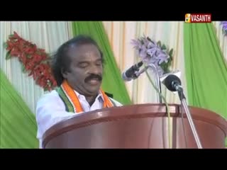 A man with a mustache and a white shirt is speaking behind a podium, a garland of orange and green around his neck. The Vasanth TV logo is visible in the top right corner.
