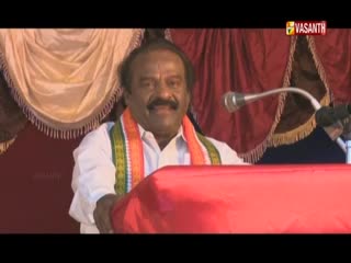 A man in a white shirt and a colorful scarf stands at a podium, speaking into a microphone. The Vasanth TV logo is visible in the corner, and red curtains form the backdrop.
