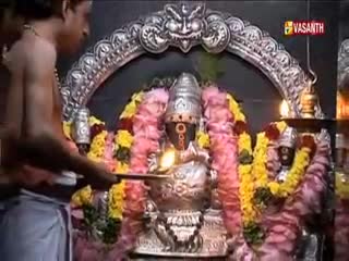 A man in a white dhoti holds a small lamp, its flame illuminating a shrine. The Vasanth TV logo is visible as the camera captures the ritual.
