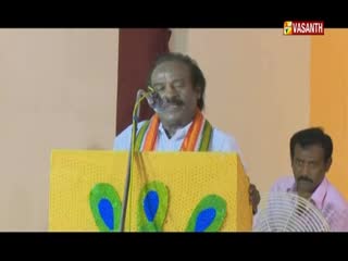 A man in a white shirt and orange scarf speaks from behind a yellow podium decorated with peacock feathers.  Beside him, another man sits, holding a fan, as the Vasanth TV logo flashes in the corner.
