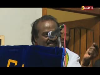 A man speaks into a microphone, his expression animated as he stands behind a blue podium. The Vasanth TV logo is visible in the corner, suggesting a broadcast from India.

