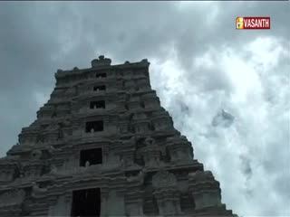 A towering temple structure rises towards the overcast sky. The Vasanth TV logo sits in the corner as the stone edifice pierces the clouds.
