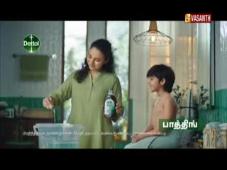 A woman in a green shirt pours liquid from a Dettol bottle into a bucket, while a young boy sits nearby. The scene is part of a Vasanth TV commercial, likely from India.
