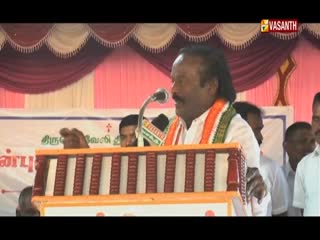 A man in a white shirt and an orange scarf speaks into a microphone from behind a podium. The Vasanth TV logo is visible in the top right corner, and a banner with Tamil script is behind the speaker.
