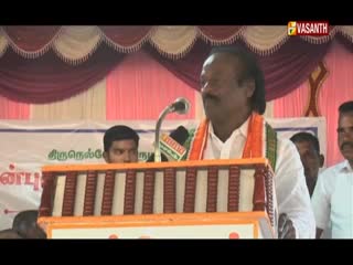 A man in a white shirt, with an orange scarf, speaks into a microphone from behind a podium. The Vasanth TV logo is visible in the corner, and a banner with Tamil script is behind the speaker.
