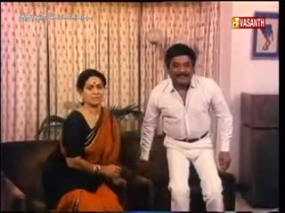 A woman in a vibrant orange sari sits on a sofa, while a man in a white shirt and pants gestures with his hands. The Vasanth TV logo is visible in the corner, suggesting this is a scene from a television program in India.

