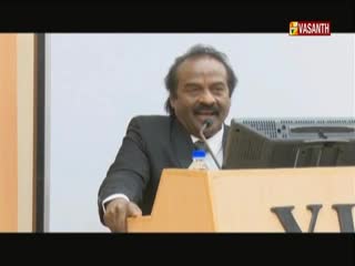 The man in the suit is speaking into a microphone, his hand resting on the podium. Behind him, the Vasanth TV logo is visible, and a computer monitor sits on the desk.
