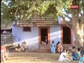 People are gathered outside a simple house, some sitting on the ground and others standing near the doorway. The Vasanth TV logo is visible in the corner, suggesting this might be a broadcast from India.
