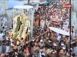 A large, ornate structure is being carried through a dense crowd. Above, a Vasanth TV logo marks the scene as a broadcast from India.
