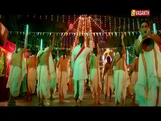 A group of men in white dhotis and kurtas are dancing, some with their arms raised. The Vasanth TV logo is visible, and the scene is lit with festive lights.
