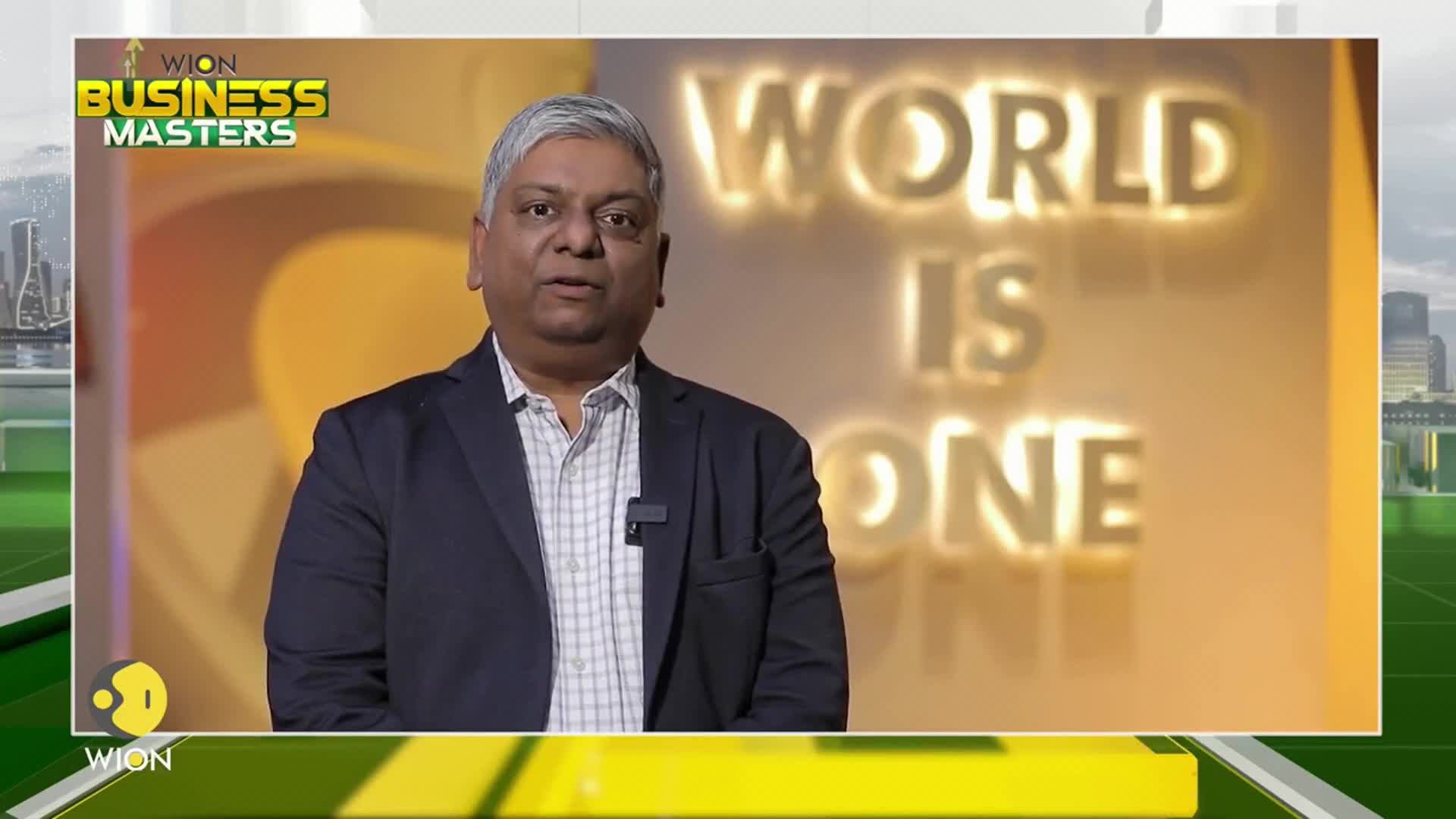 A man in a blazer speaks directly to the camera, a small microphone clipped to his lapel. Behind him, the words "WORLD IS ONE" glow against a warm, golden backdrop.
