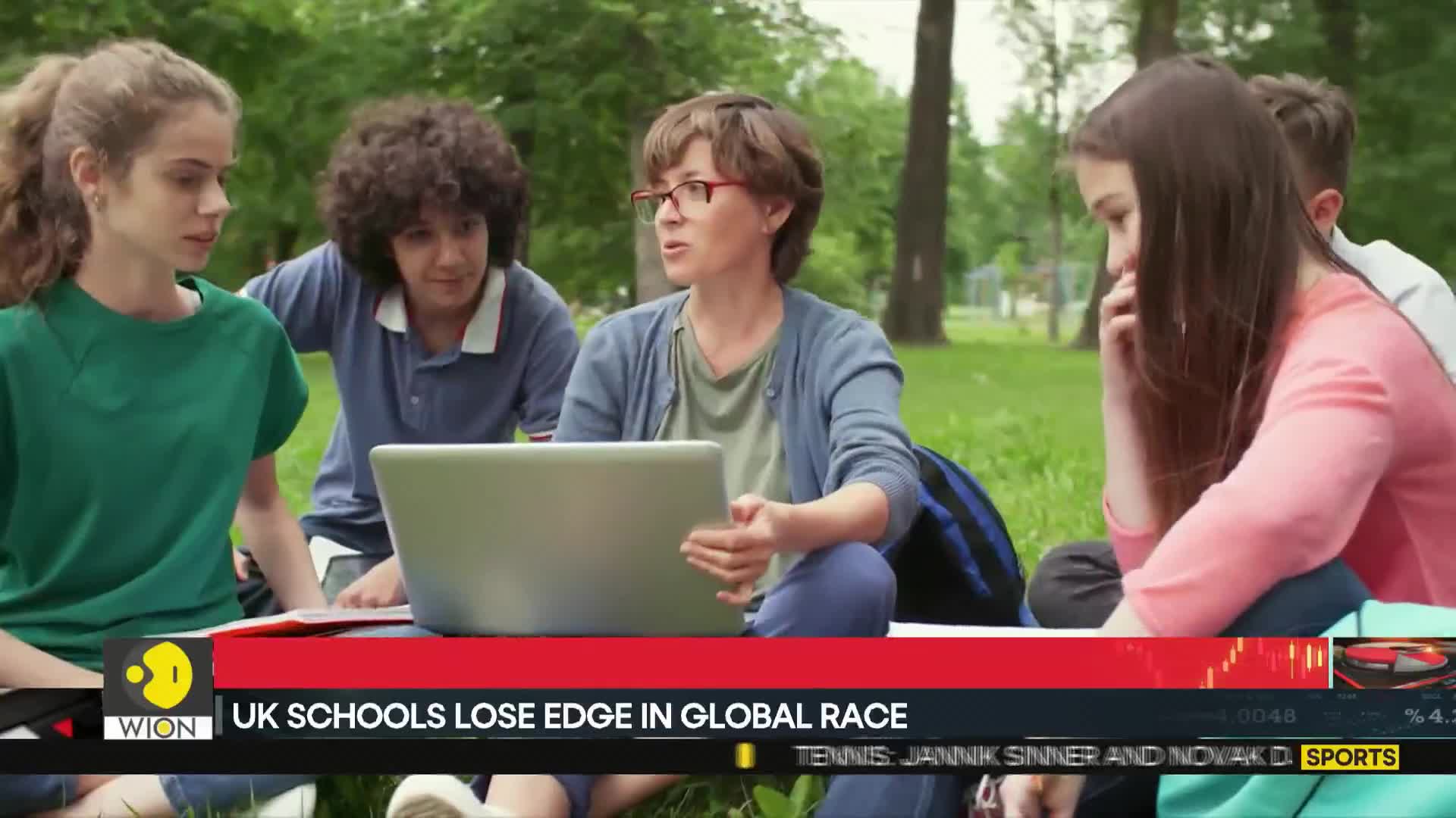 A woman with glasses gestures while speaking, a laptop open in her lap.  Four students listen attentively, seated around her on the grass.
