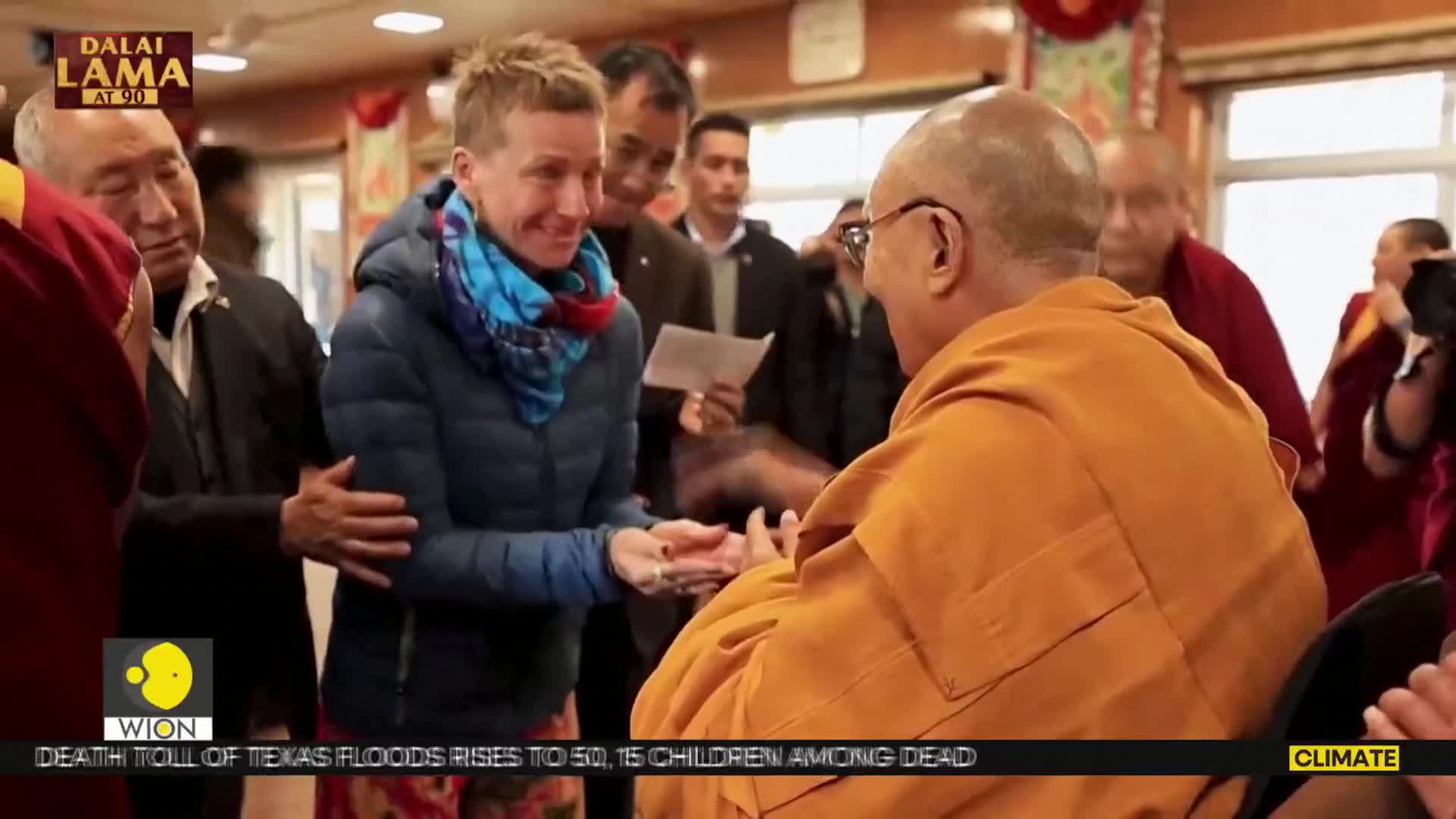 A woman in a blue jacket and scarf reaches out to the Dalai Lama, who is seated and wearing an orange robe. Several people stand nearby, watching the interaction.
