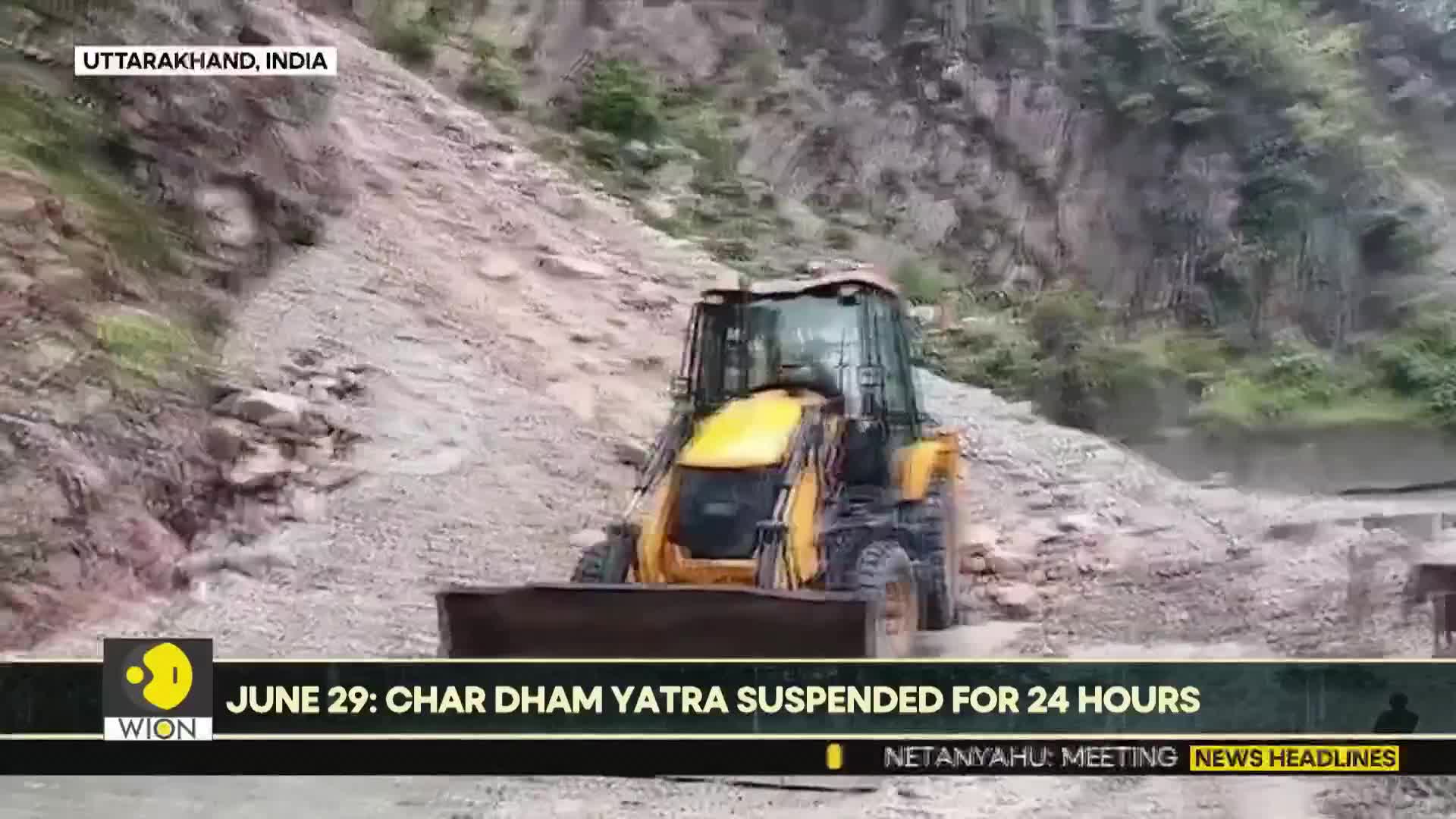 A yellow excavator is clearing debris from a hillside in Uttarakhand, India. Dust plumes rise as the machine pushes the rubble forward.
