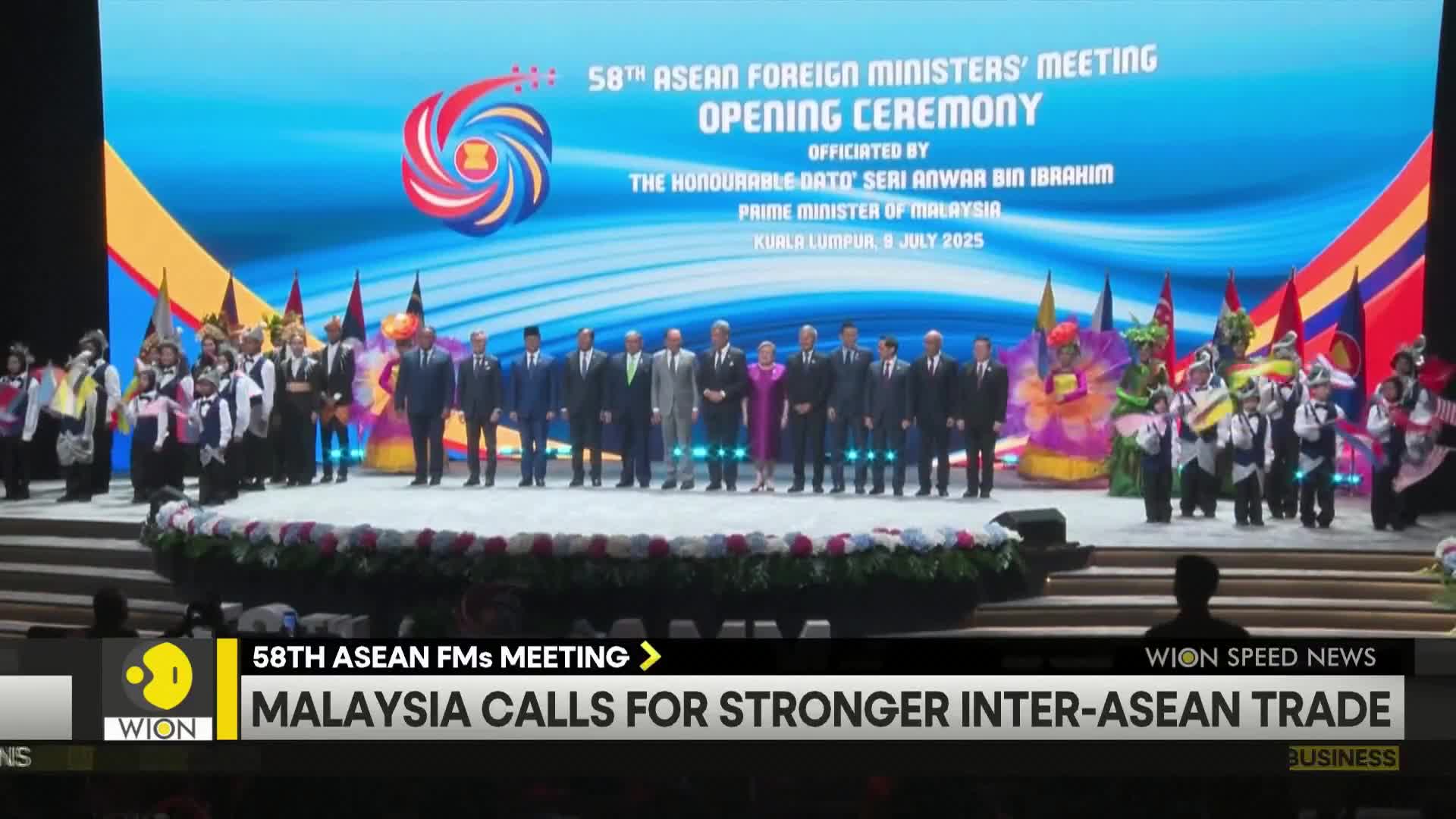 On a stage, a group of suited figures stands in front of a large screen displaying the ASEAN Foreign Ministers' Meeting. Performers in colorful costumes and flags are positioned on the left side of the stage.
