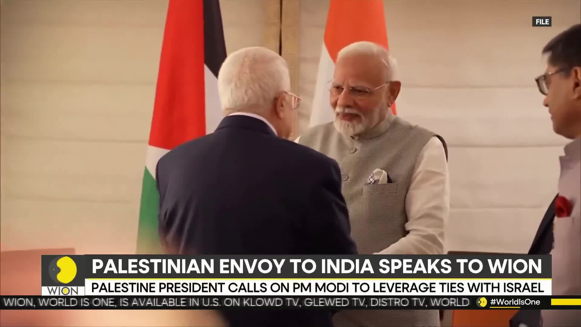 The Prime Minister of India is smiling, shaking hands with a man in a dark suit. Behind them, the flags of Palestine and India stand side-by-side.
