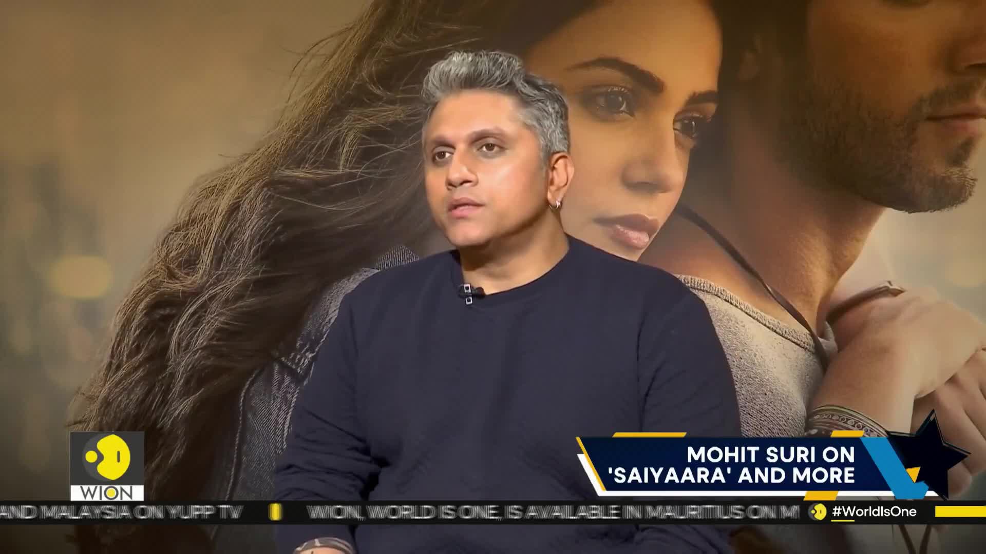 A man with silver hair, wearing a dark sweater, looks intently at someone off-camera. Behind him, a movie poster featuring a couple is slightly visible, and the WION logo is in the corner.
