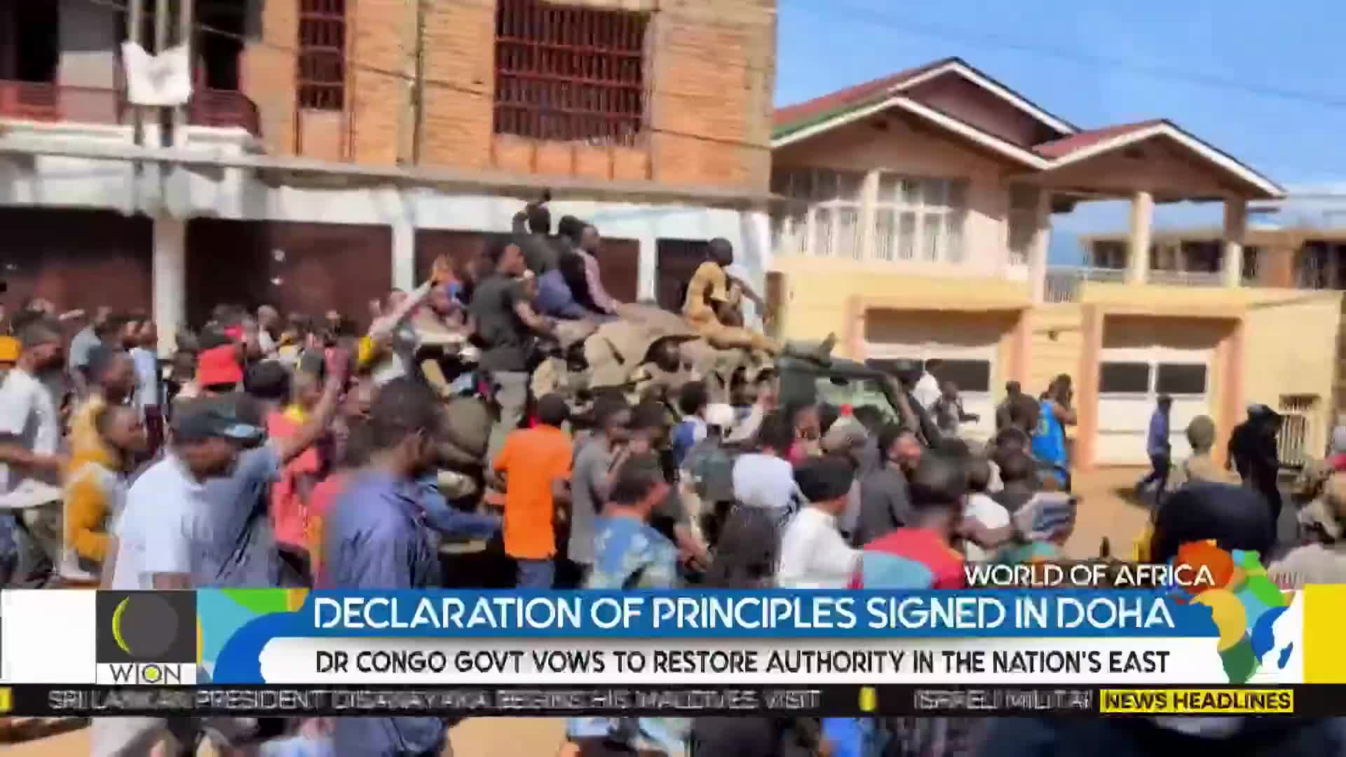 A crowd surrounds a military vehicle, with people climbing on top and waving. The scene unfolds in front of buildings, with a news banner from WION at the bottom.
