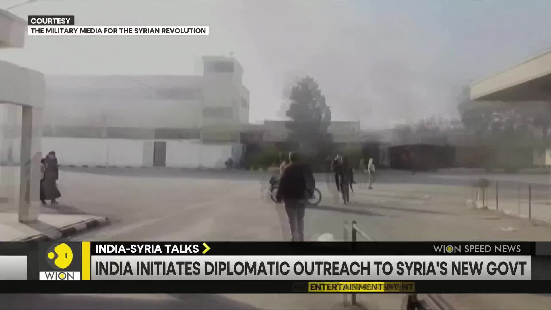 A few people walk across a large, mostly empty paved area, some heading towards buildings in the distance. The scene is hazy, with a WION news banner at the bottom reporting on India's diplomatic outreach to Syria.
