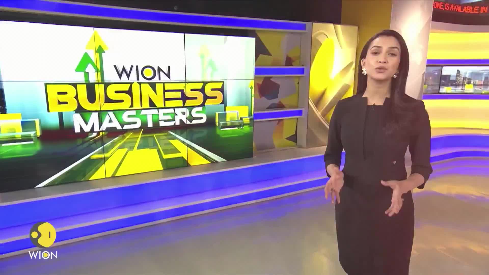 A woman in a black dress gestures with her hands in front of a large screen displaying the "WION Business Masters" logo. The set is brightly lit with yellow and blue accents.
