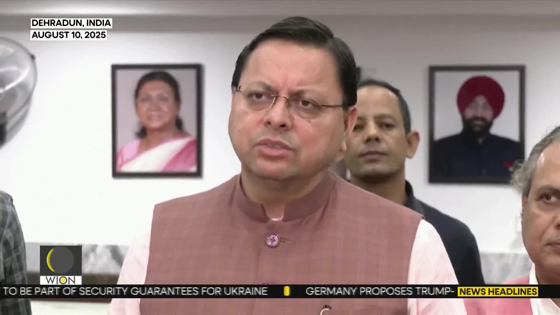 A man in a pink vest speaks, flanked by other men and framed portraits, as a news ticker from WION scrolls at the bottom. The broadcast is originating from Dehradun, India.
