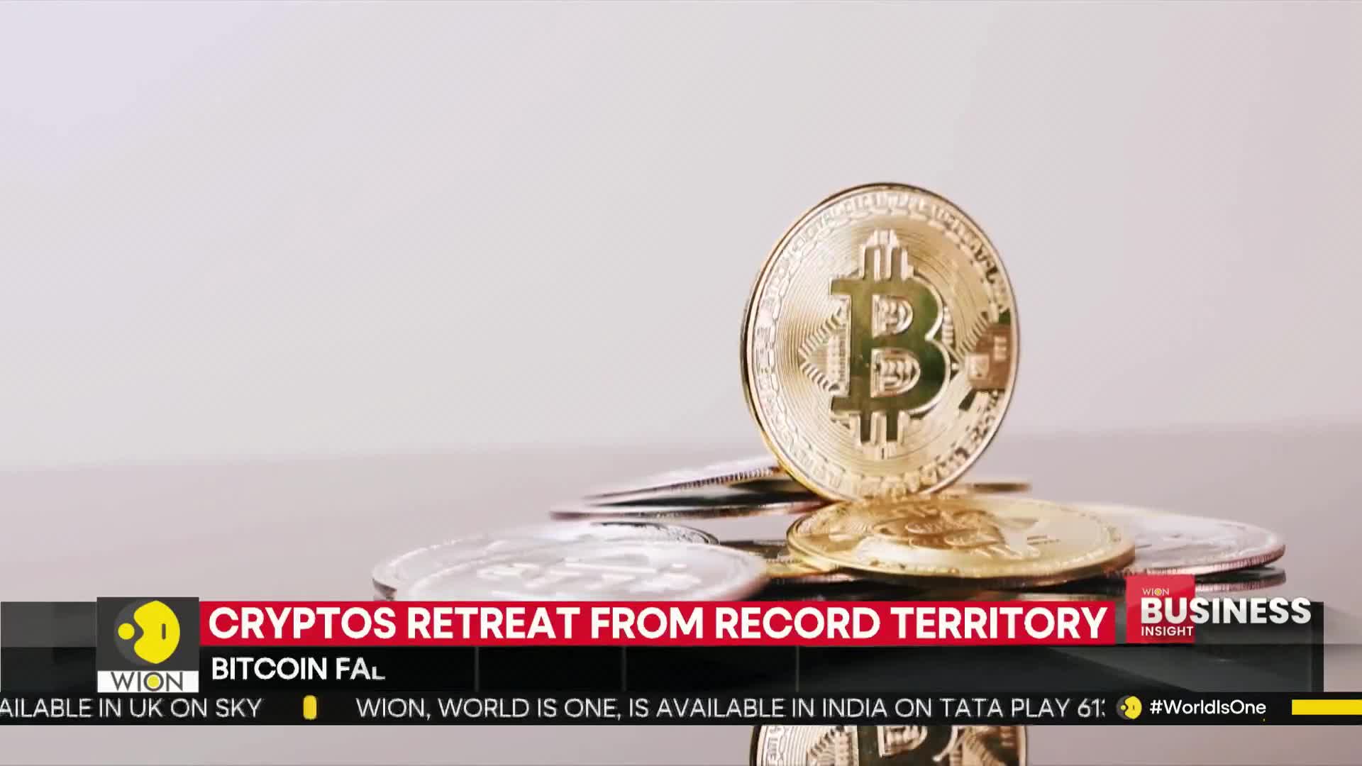 A large gold Bitcoin coin stands upright, leaning against a small pile of other coins. Below, a WION news graphic from India announces a decline in crypto value.
