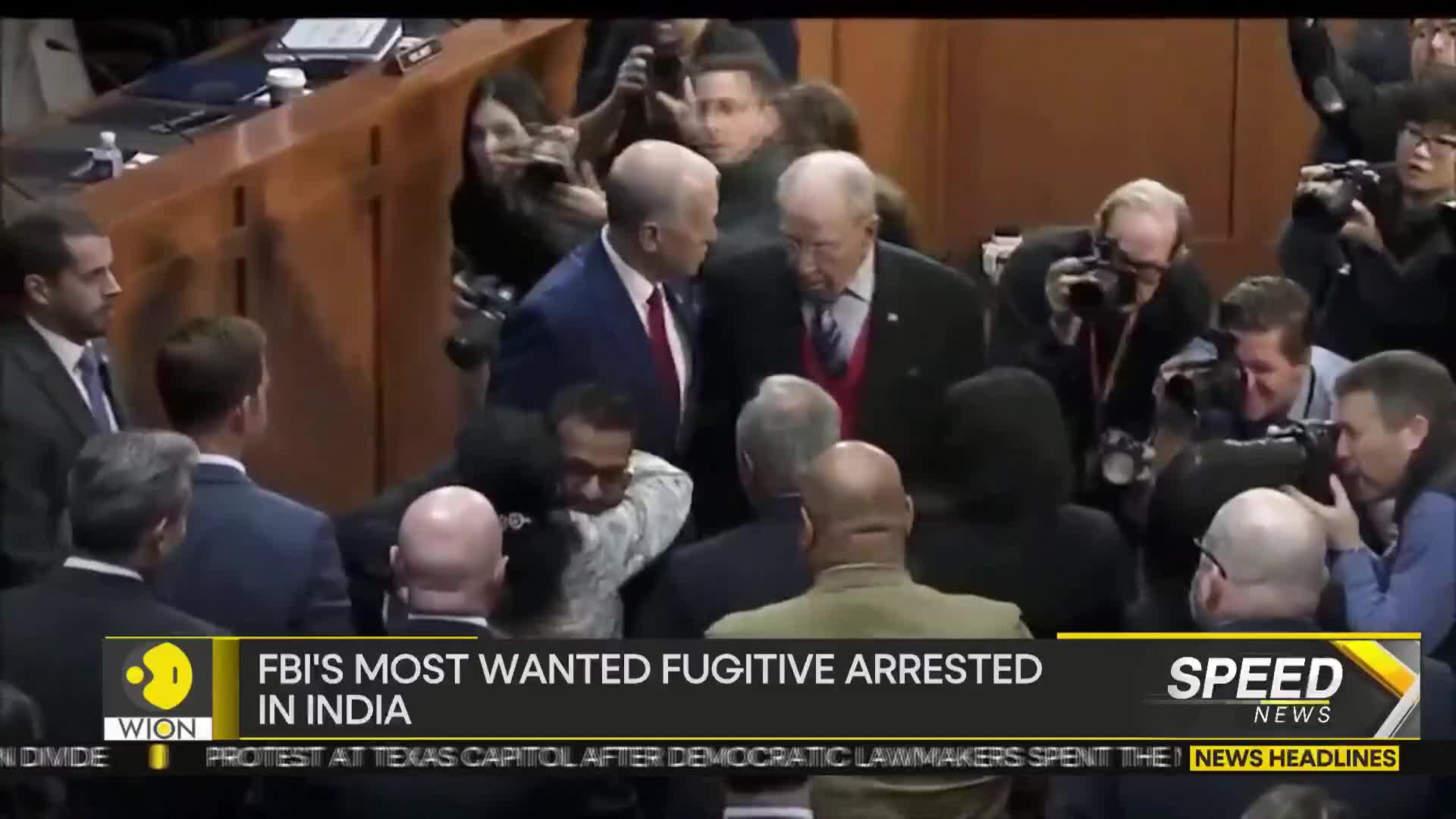 A group of men in suits are surrounded by people holding cameras, some snapping photos. The WION news banner at the bottom of the screen reads "FBI's Most Wanted Fugitive Arrested in India."
