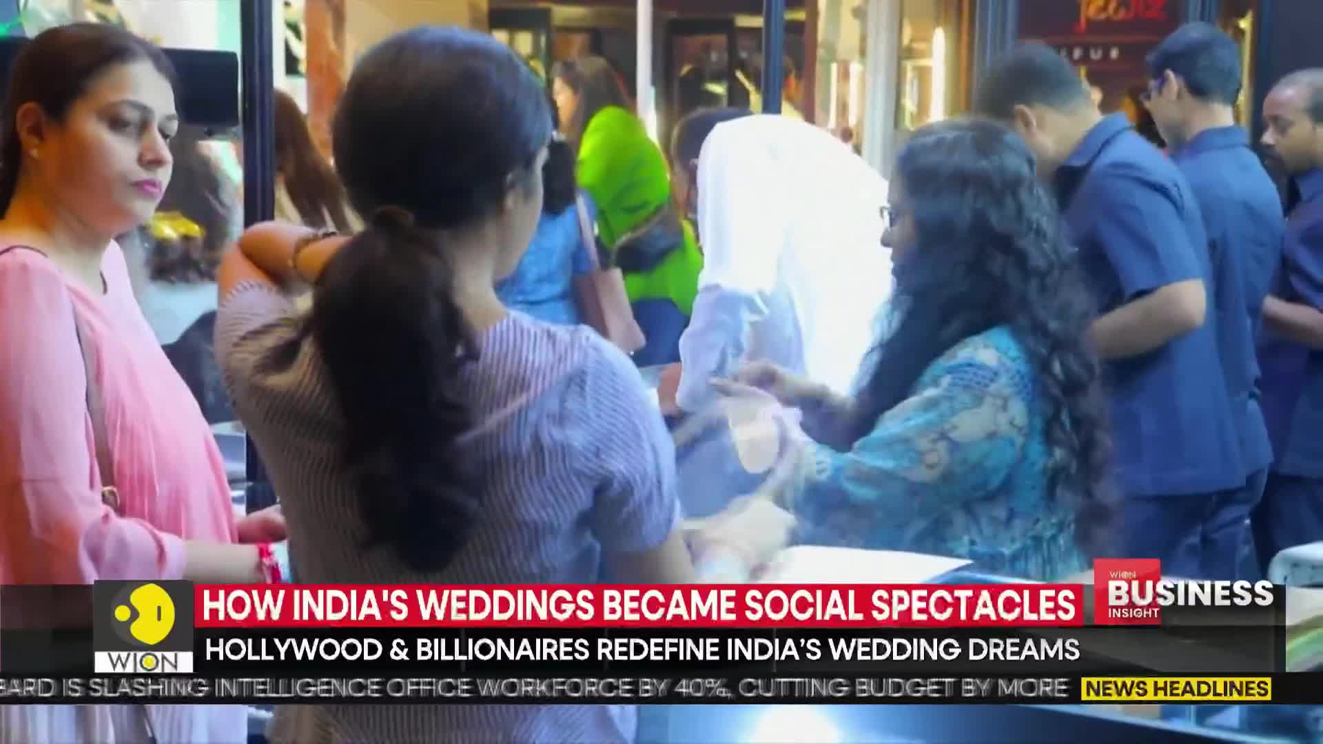 People are gathered, some looking at something out of view, while others observe. The scene appears to be in a shop or a similar setting, with a news headline about Indian weddings displayed on the screen.
