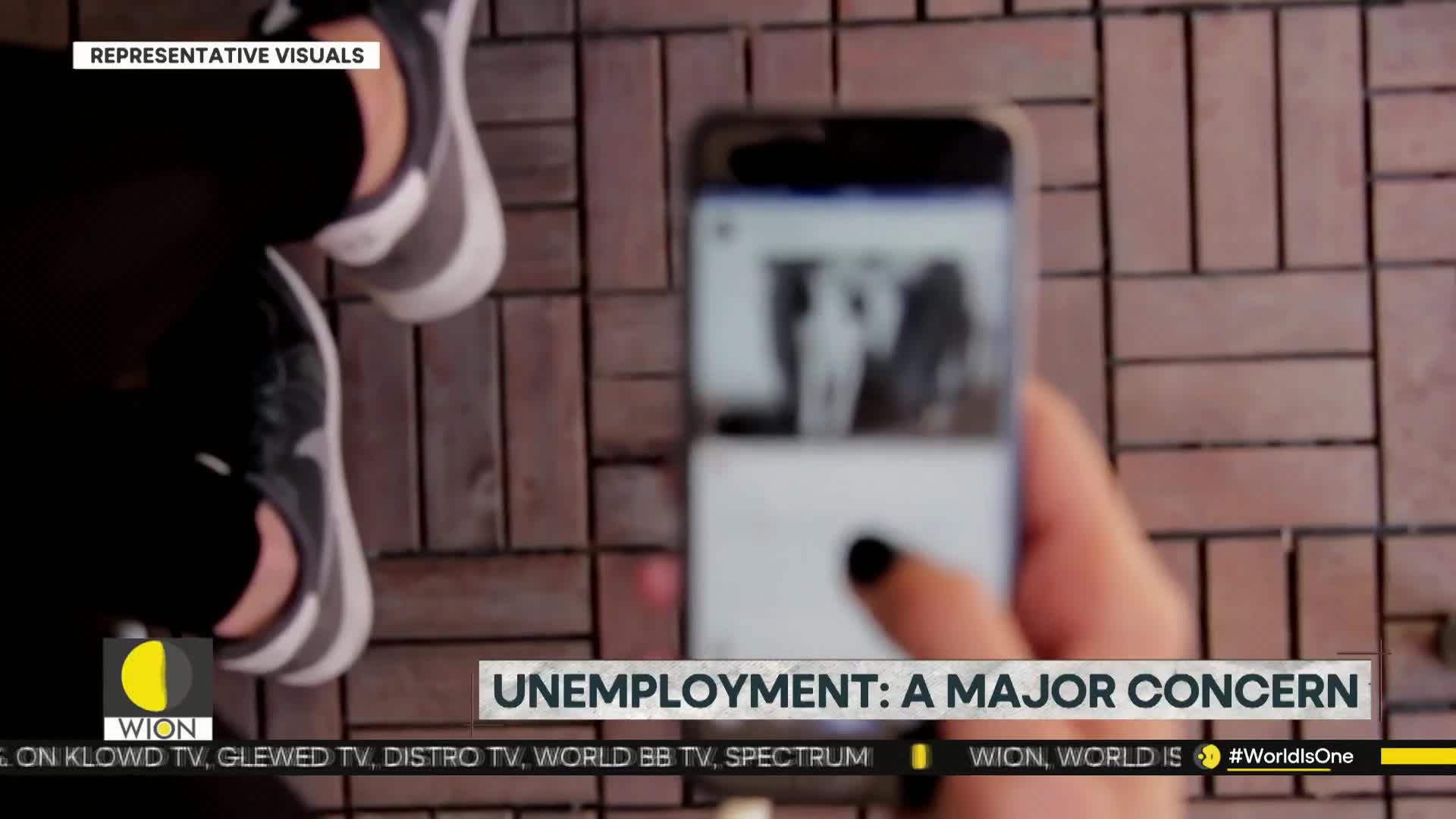 A hand with a black-polished fingernail taps a phone screen, the image on it slightly out of focus. The phone is held over a brick-patterned surface, and the person's feet in sneakers are visible in the background.
