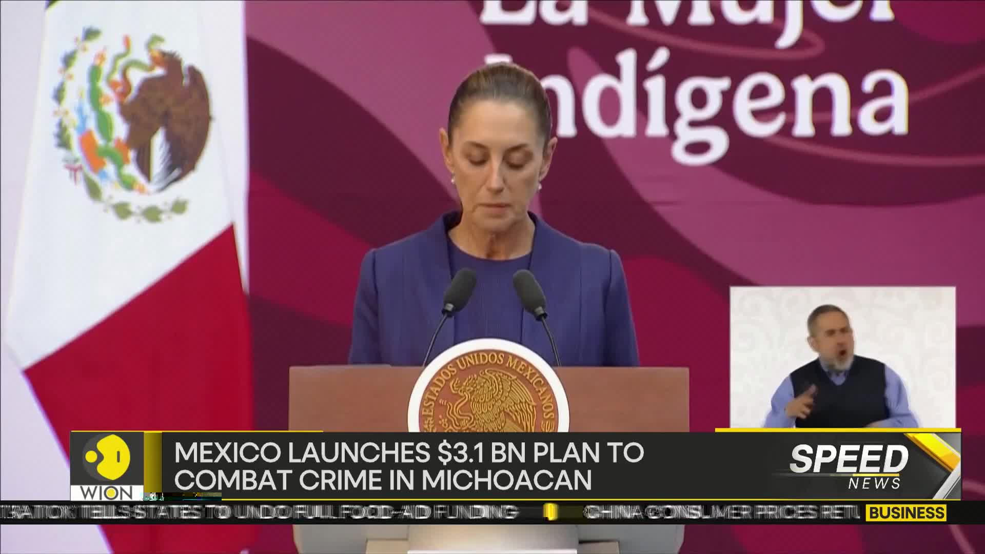 A woman in a blue suit stands at a podium, speaking into two microphones. The Mexican flag is visible behind her, and a sign language interpreter is in a small box on the side.
