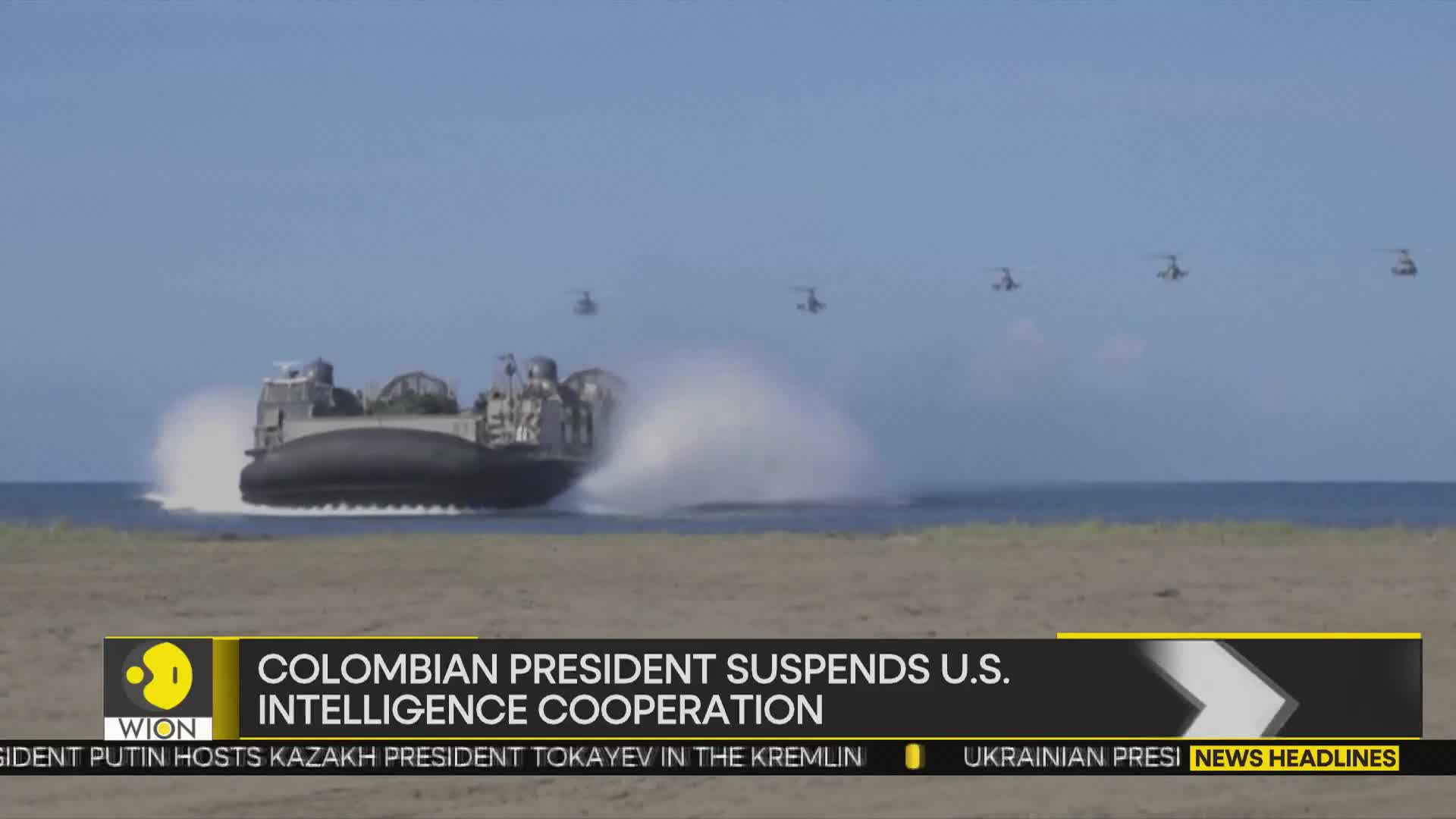 A large, dark hovercraft is skimming across the water, kicking up a huge spray. Above, a line of helicopters flies in formation against a clear blue sky.
