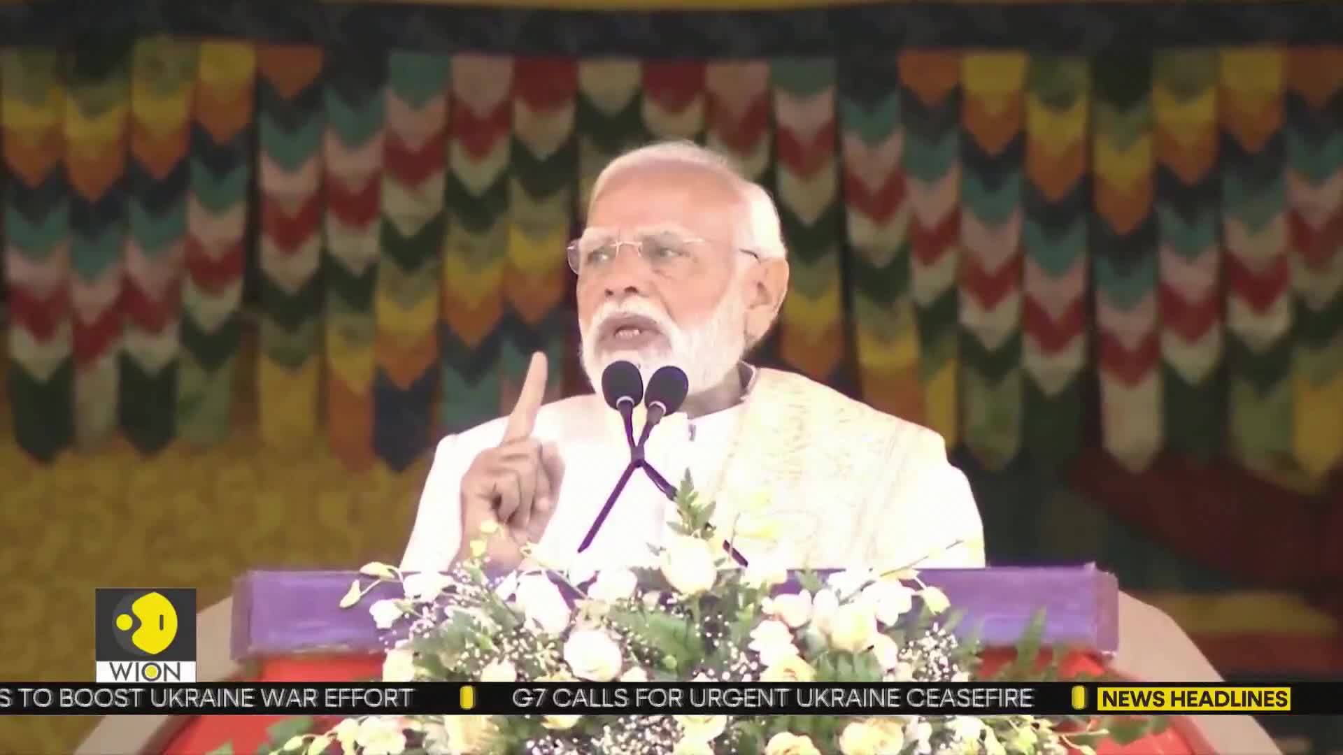 A man in a cream-colored garment gestures with his right index finger while speaking into two microphones. Behind him, a colorful backdrop hangs above a stage decorated with white flowers.
