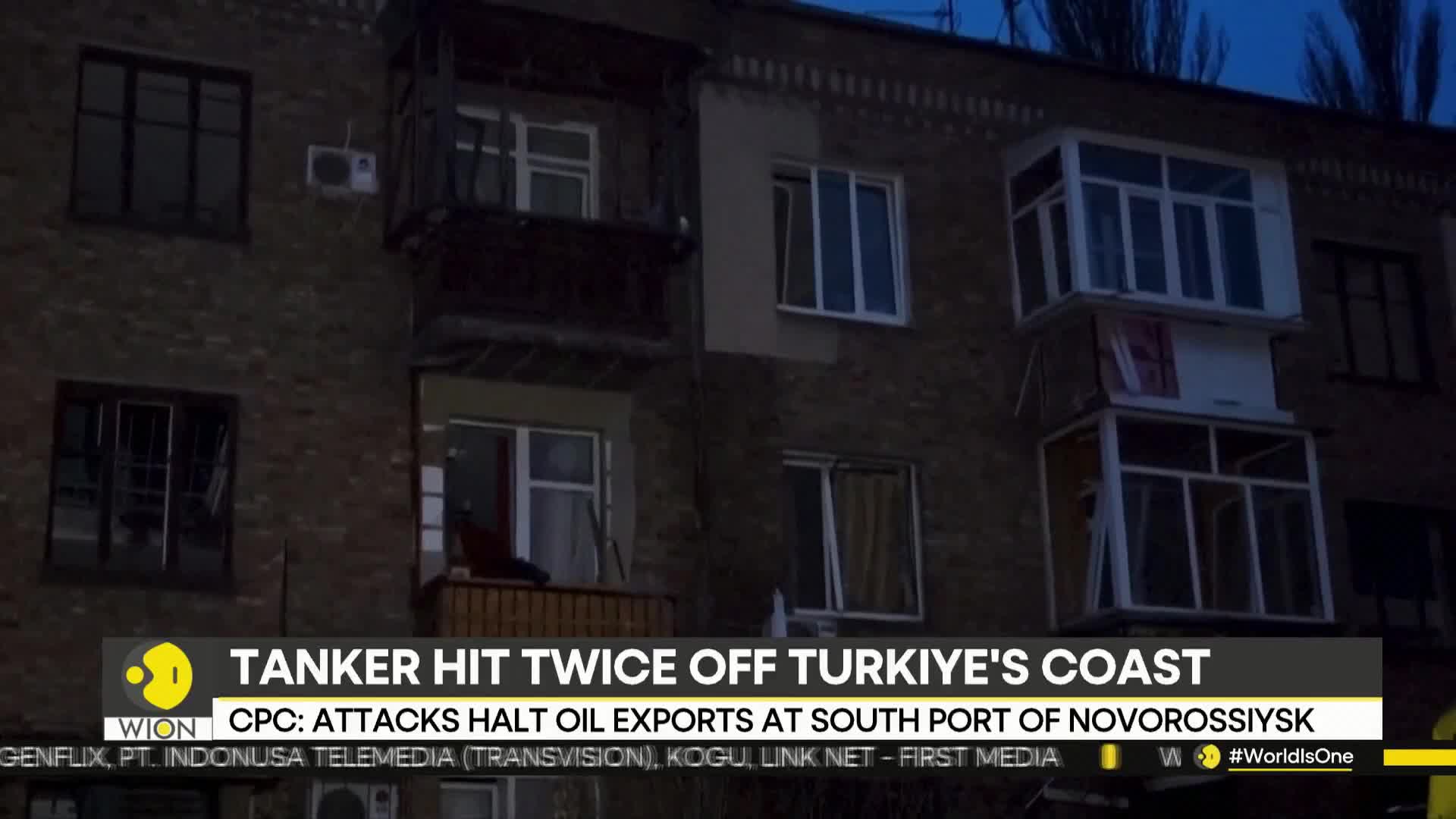 Darkness has fallen on a drab apartment building, its windows reflecting the dim light. A news ticker from WION scrolls at the bottom, reporting on attacks and oil exports.
