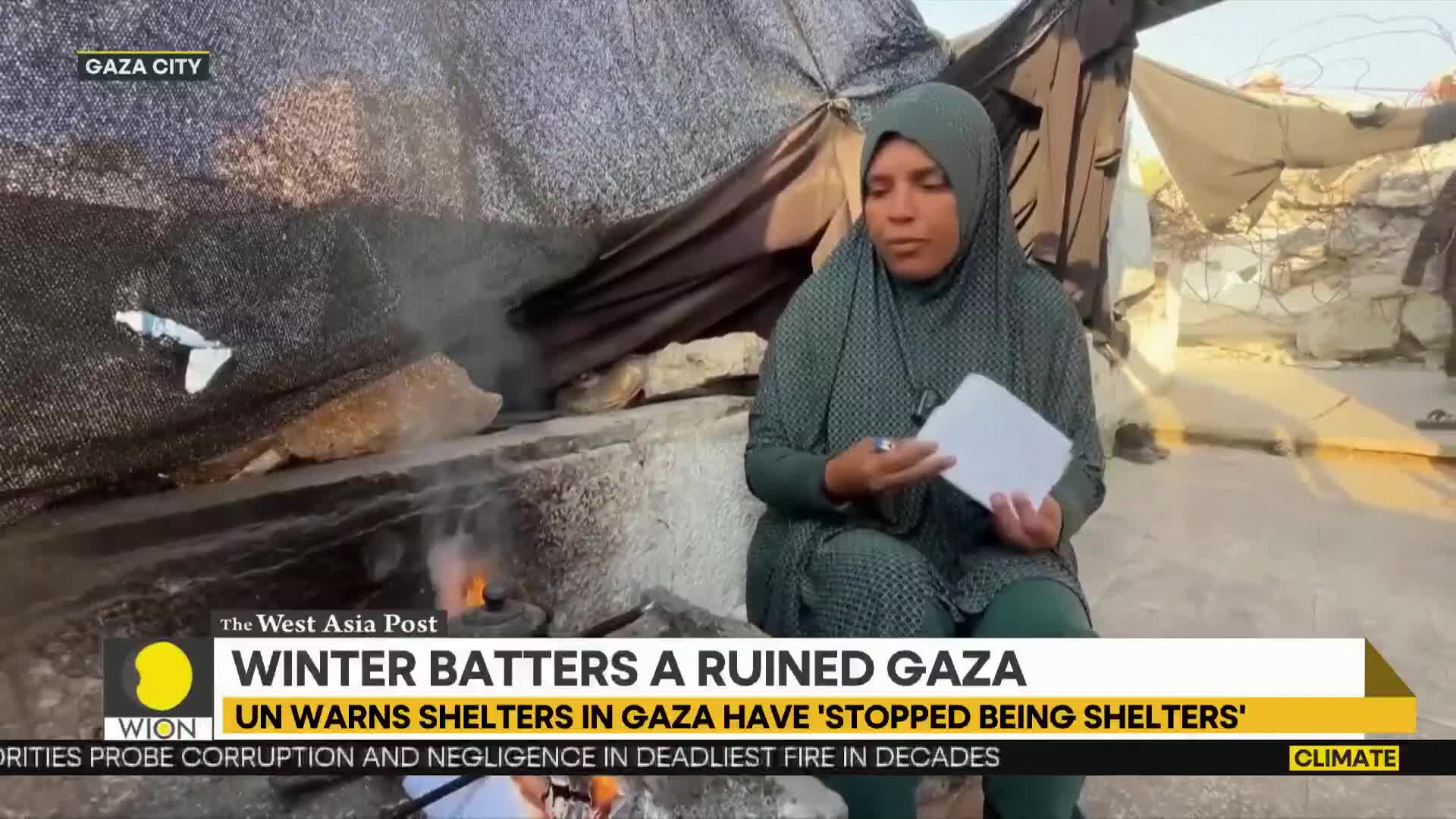 A woman in a hijab sits near a small fire, holding papers and speaking. Smoke drifts upwards from the flames, and the backdrop suggests a damaged structure.
