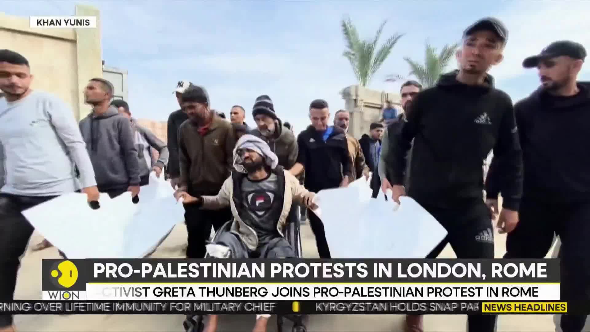 A group of men are walking forward, some carrying large white sheets. A man in a wheelchair is being pushed along with them, his hands open as if addressing the crowd.
