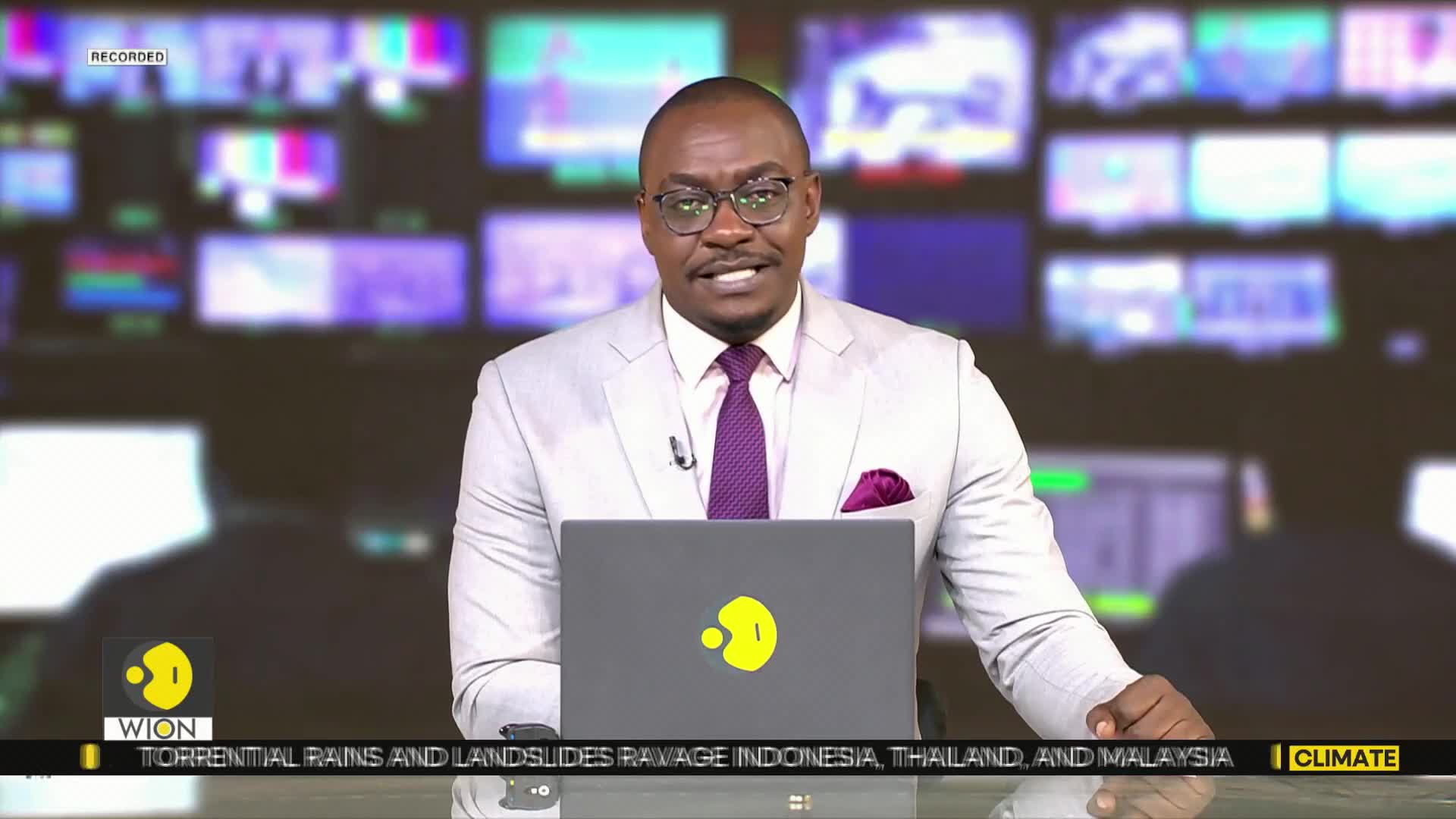 A man in a light suit and glasses speaks directly into the camera, a laptop open in front of him. Behind him, a wall of screens displays various images, and the WION logo is visible on the lower left.
