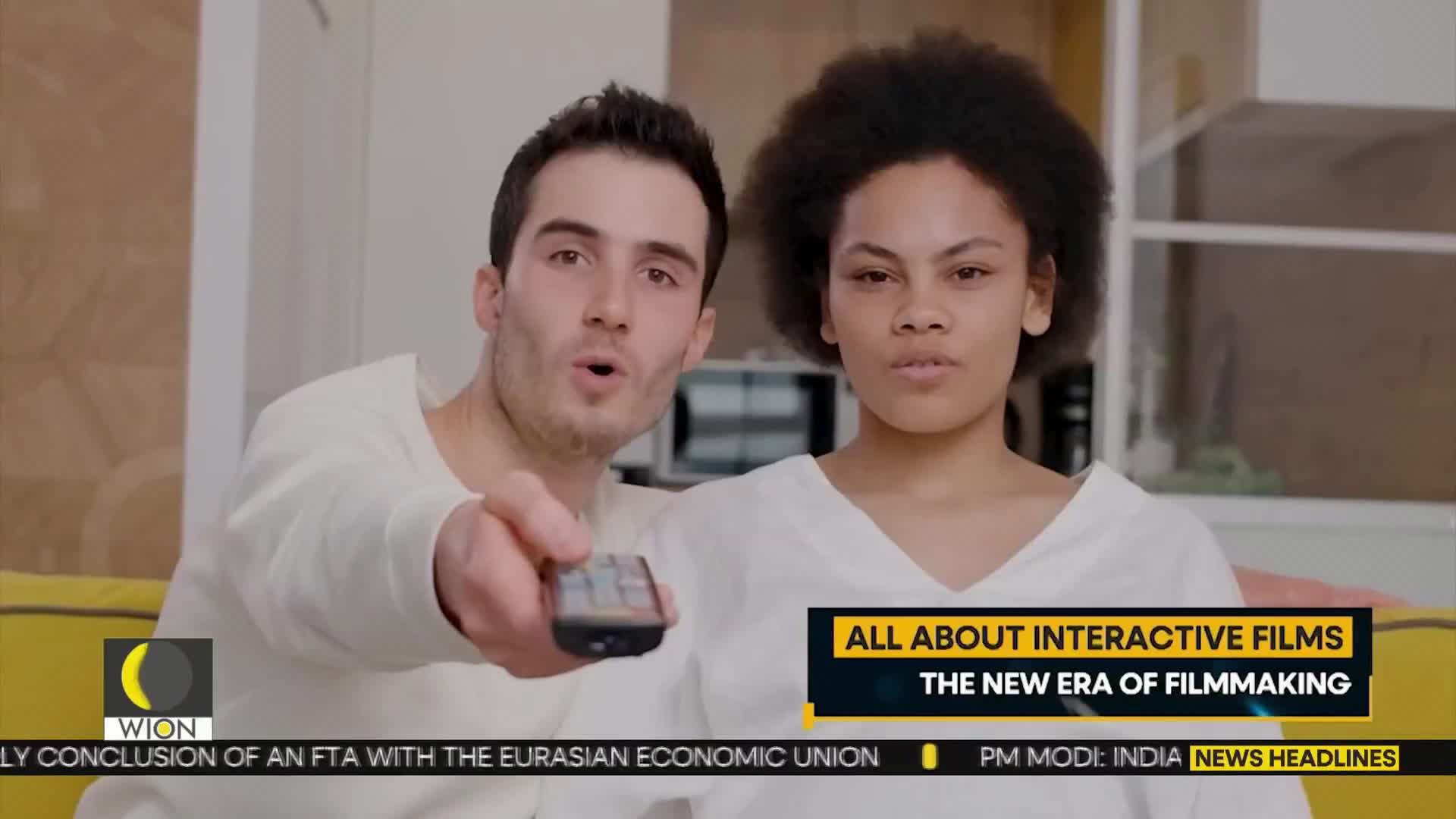 A man, holding a remote, points it towards the screen while his companion watches intently. The WION logo and news headlines are visible at the bottom of the screen.
