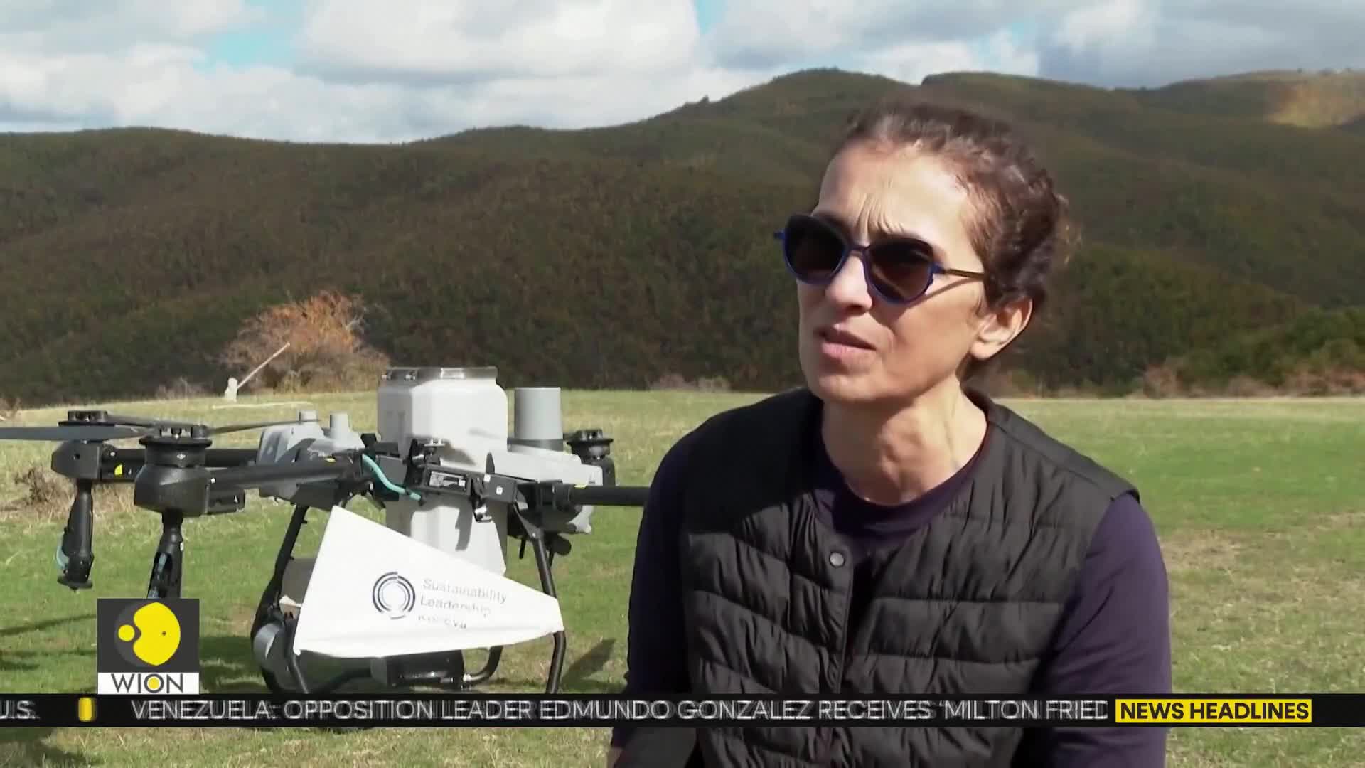A woman wearing sunglasses is speaking, a drone with a white banner behind her. The WION logo is in the corner, and the backdrop is a green field with hills.
