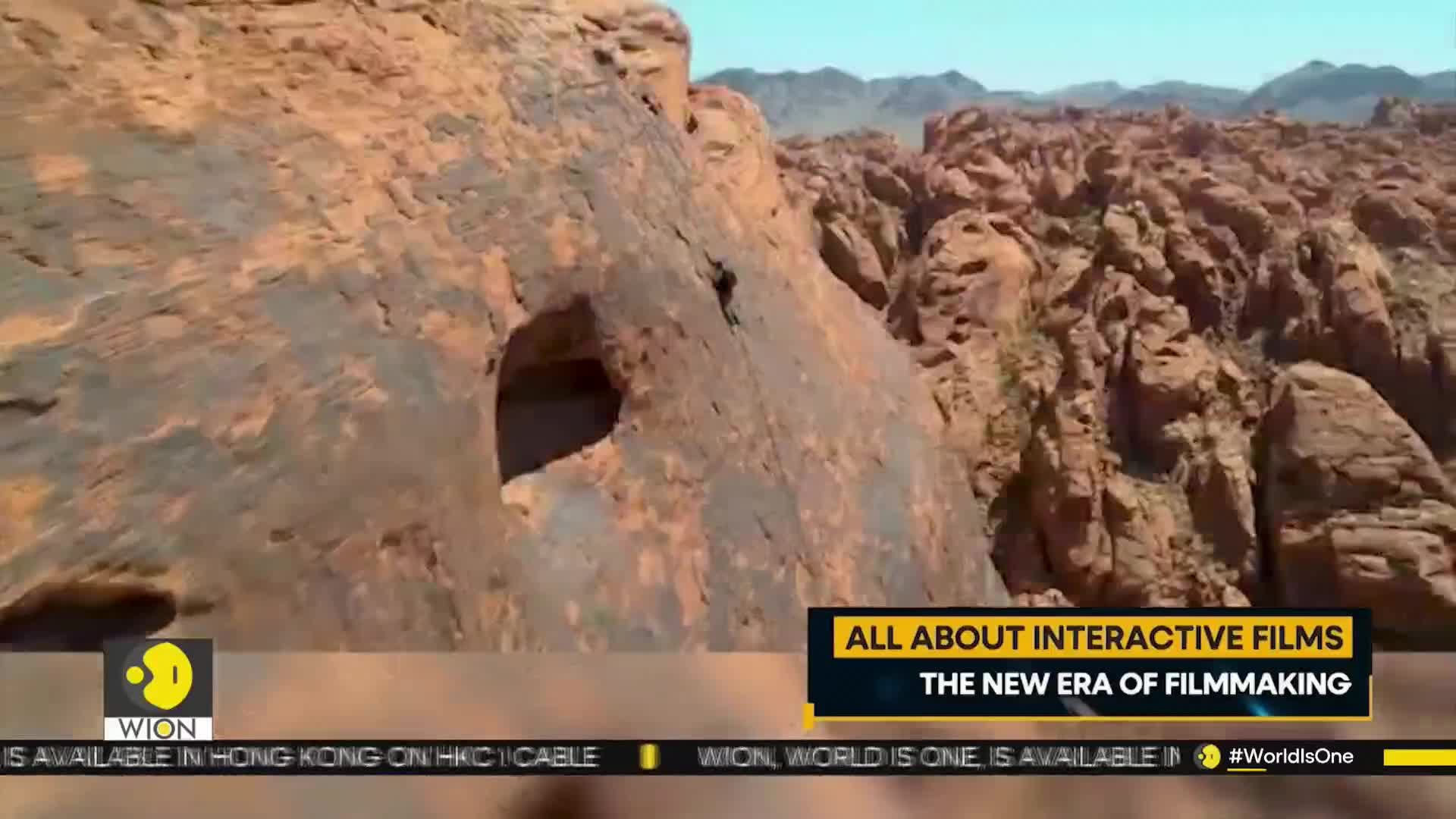 A person, tiny against the rock face, is climbing near a hole in the sandstone. The scene, broadcast by WION, shows a vast, textured landscape of red rock formations.
