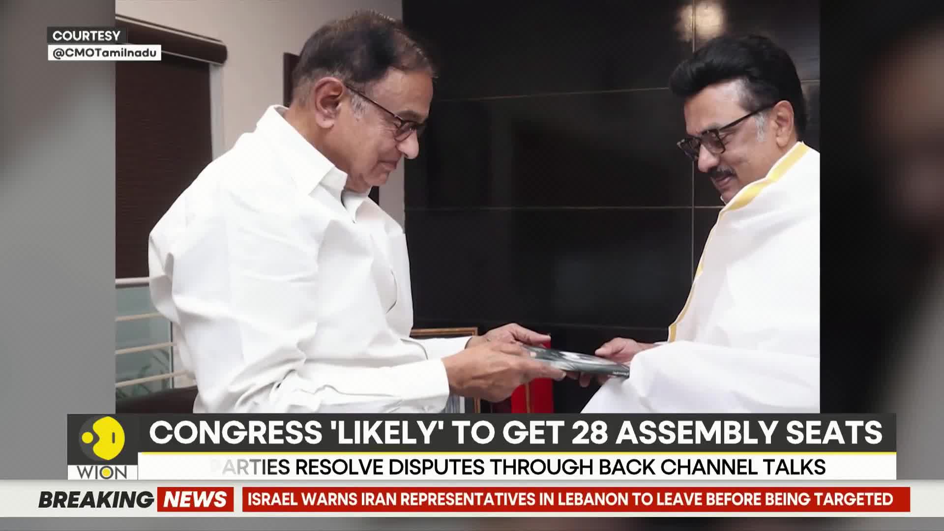 Two men in white shirts exchange papers. The one on the left, wearing glasses, hands a document to the man on the right, who is draped in a white shawl.