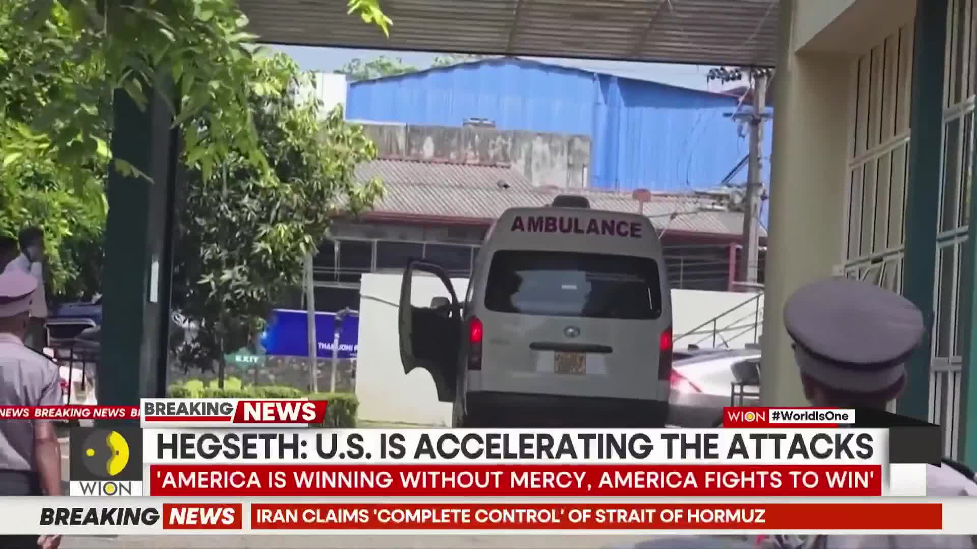 An ambulance with its doors open backs up to a building. Two men in uniforms stand nearby, watching.