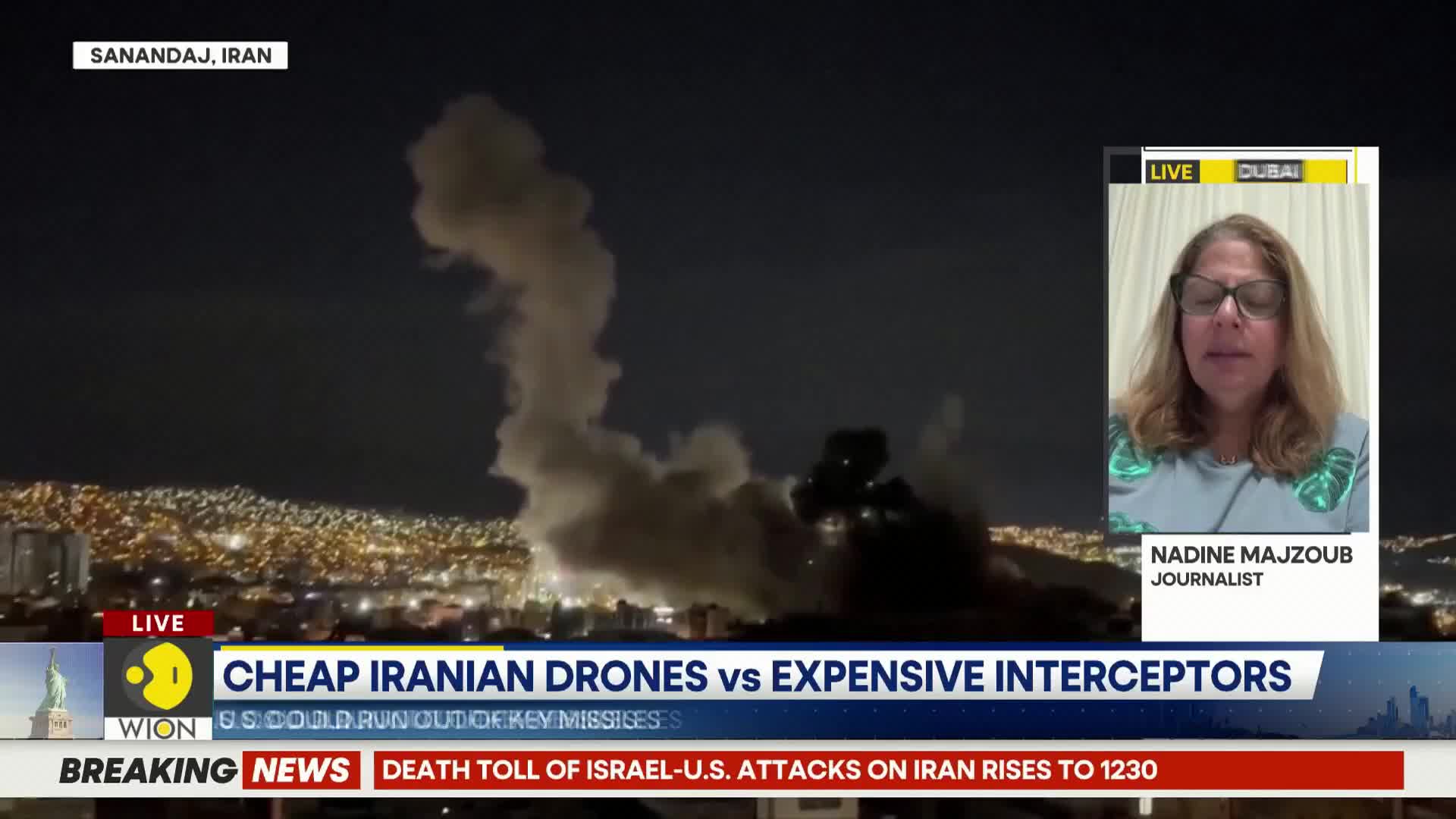 Thick plumes of smoke billow into the night sky over Sanandaj, Iran, illuminated by the city's lights. Explosions erupt in the distance, sending sparks and debris upwards.